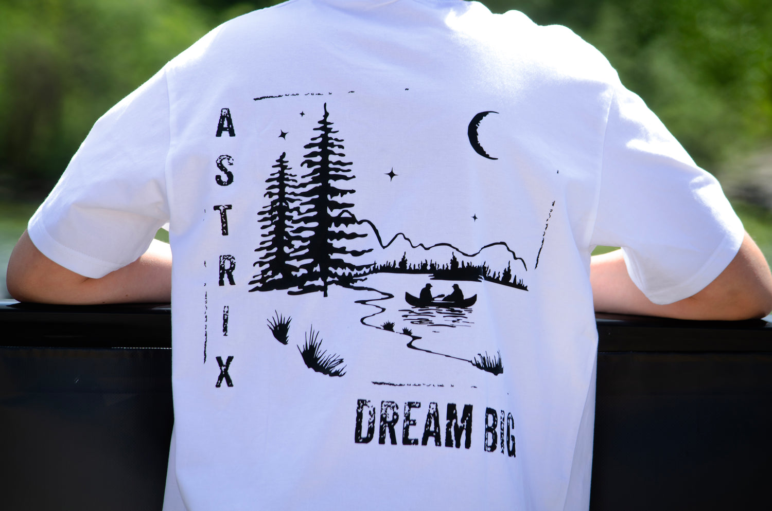 White t-shirt with black nature-themed design and 'Dream Big' text on a blurred natural background